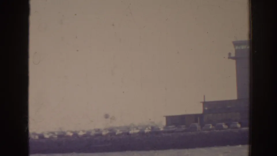 1962 Florida 8mm frame with a distant airport building, partial control tower, parked cars, and hazy sky.