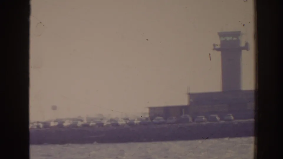 1962 Florida 8mm frame showing the airport control tower, terminal structures, parked cars, and water foreground.