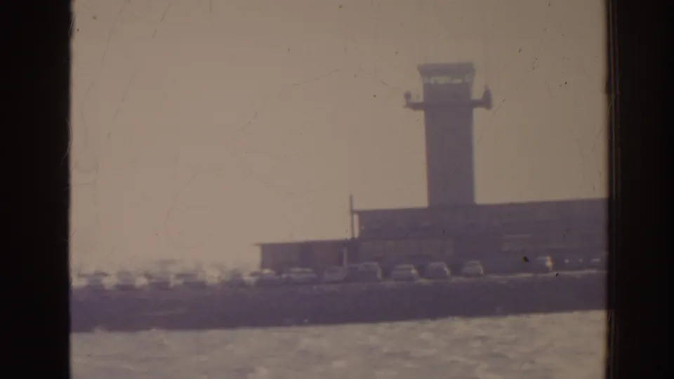 1962 Florida 8mm frame with a control tower, low airport buildings, shoreline cars, and rippled water in front.
