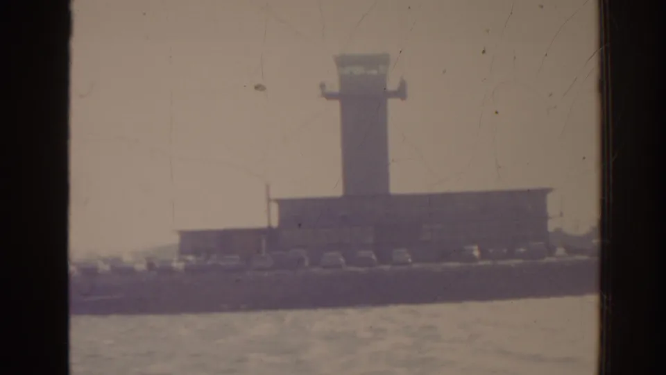 1962 Florida home-movie view of a harbor structure and waterfront through a dark vertical window frame, with hazy water in the foreground.