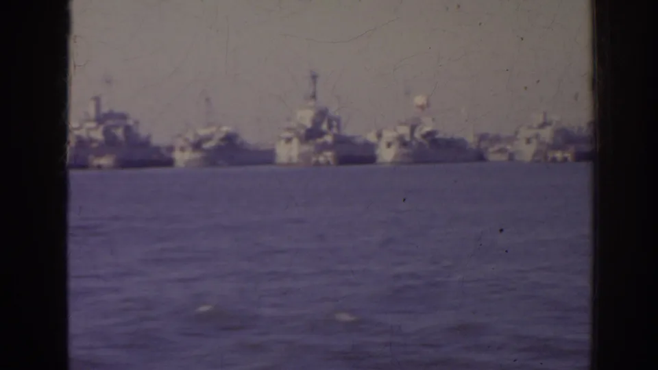 1962 Florida 8mm frame of distant naval vessels anchored across the harbor, seen through a window edge with muted purple-sepia film color.