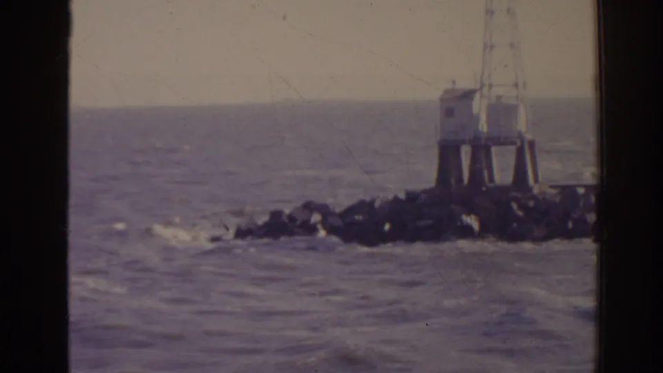 1962 Florida 8mm frame of a white beacon or lighthouse structure standing on rocks at the edge of choppy ocean water.