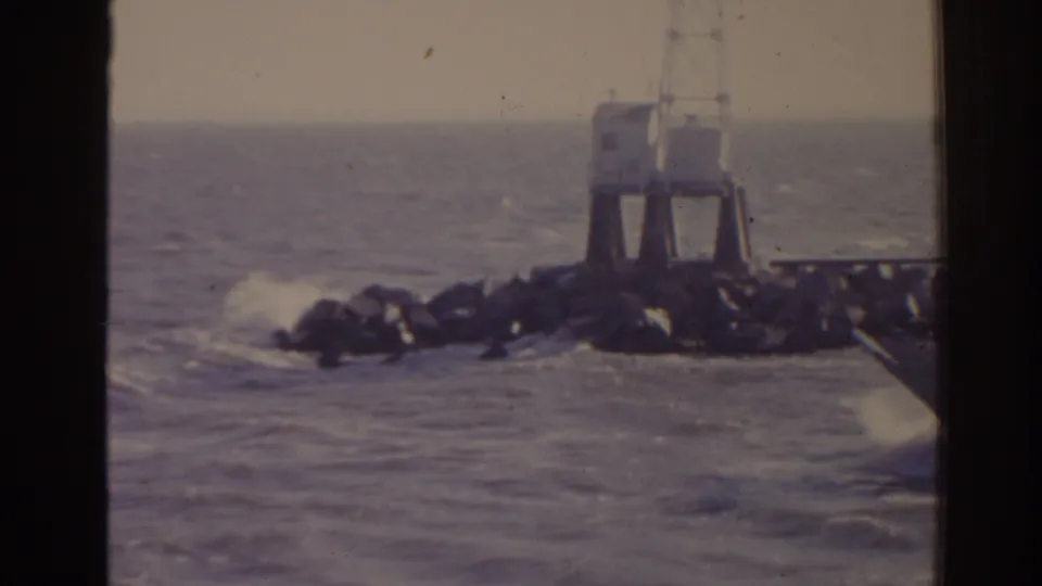 1962 Florida coastal frame showing a white lighthouse-like marker on a rocky jetty with waves breaking along the ocean edge.