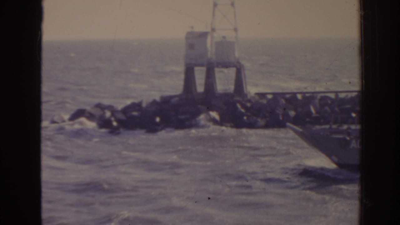 Landing craft near a rocky jetty in choppy water, seen through dark vertical frame bars.