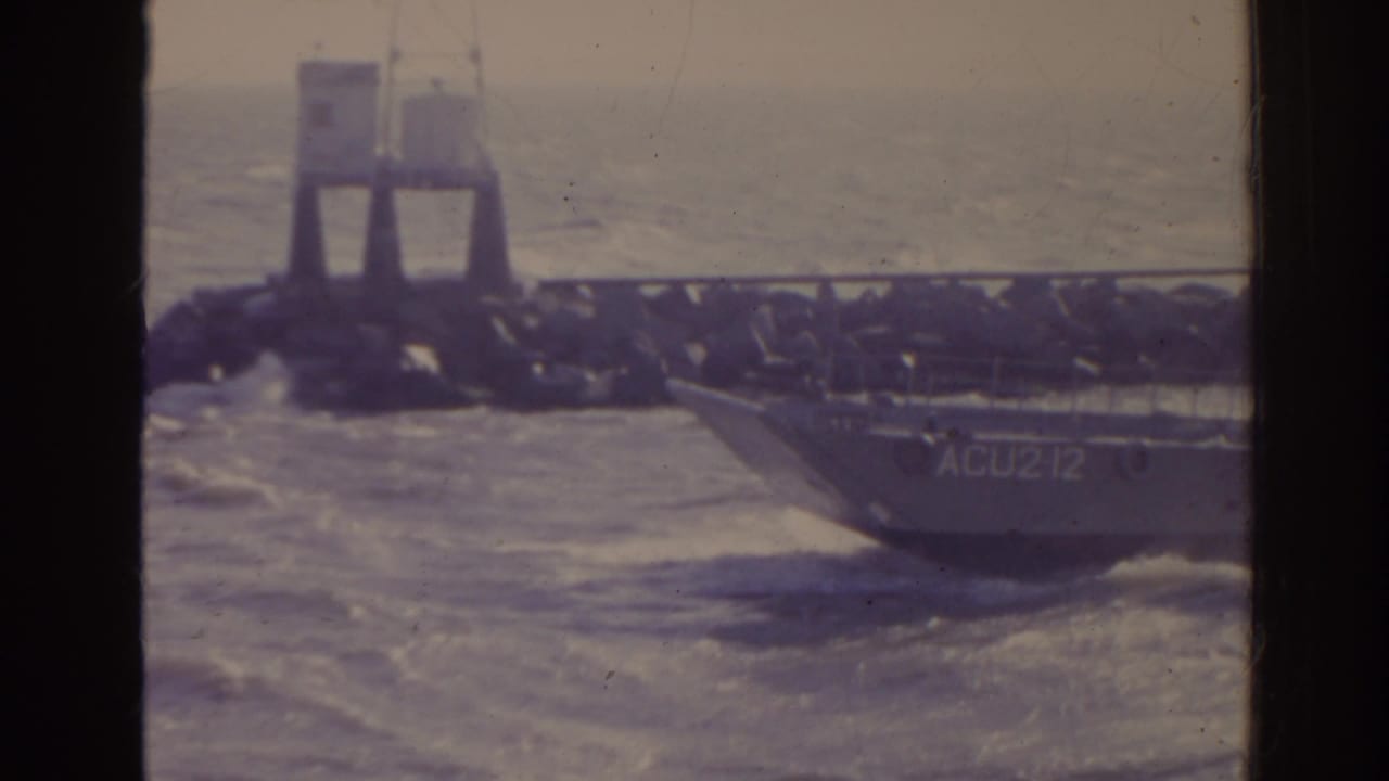 Landing craft marked ACU212 passes beside a rocky jetty in rough water.