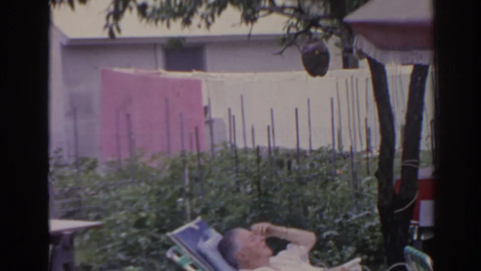1962 Florida home-movie frame of an elderly man resting in a lounge chair beneath tree shade beside a backyard clothesline.