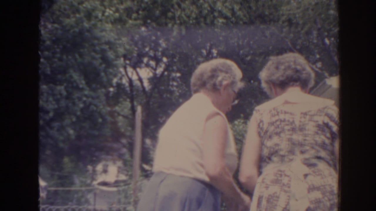 Two older women seen from behind in a leafy yard, one in a sleeveless top and skirt and one in a patterned dress.