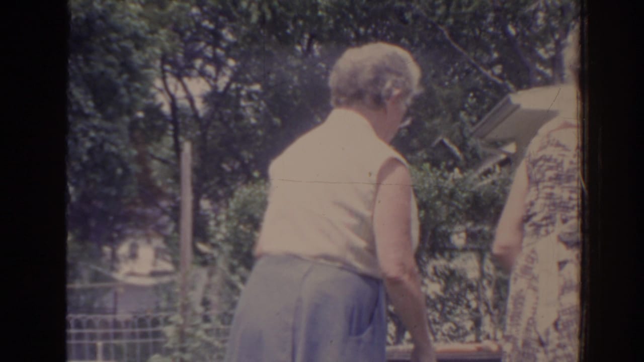 Two older women lean forward near shrubs, with trees and a fence in the background.