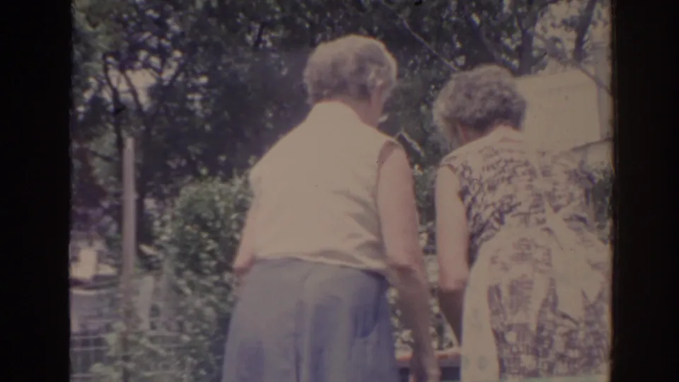 1962 Florida home-movie frame of two older women from behind, close together in a leafy yard with fencing and trees behind them.