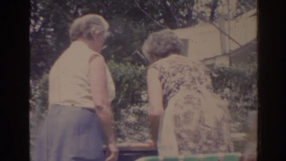 1962 Florida home-movie frame showing two older women bending in a backyard, one wearing a patterned dress with a white apron.