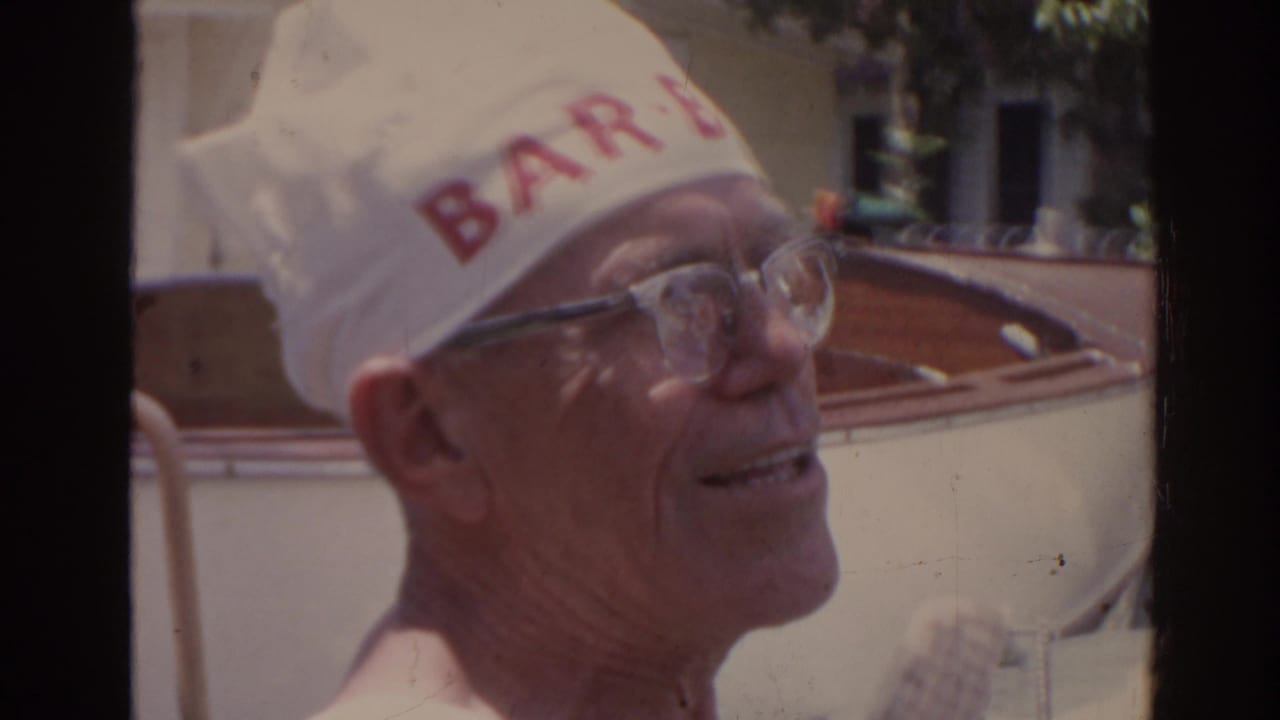 In 1962 Florida home-movie footage, an older shirtless man in glasses smiles while wearing a white cap with red lettering near a wooden boat.