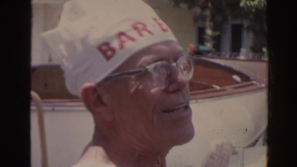In 1962 Florida home-movie footage, an older shirtless man in glasses smiles while wearing a white cap with red lettering near a wooden boat.