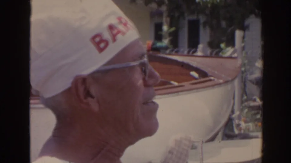 Side profile of an older man in glasses and a white cap with red lettering, with a wooden boat visible behind him in the Florida outdoor setting.