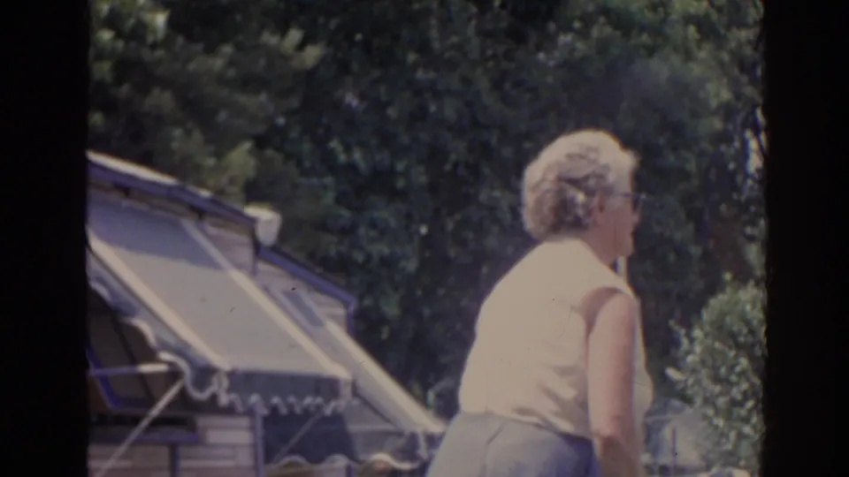 A woman seen from behind in the same 1962 Florida outdoor home-movie scene, standing near trees, shrubs, and a striped awning.