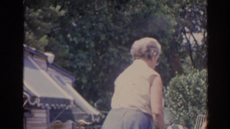 The woman continues through the Florida yard scene, seen from behind near shrubs, trees, and a striped awning in soft 8mm color.