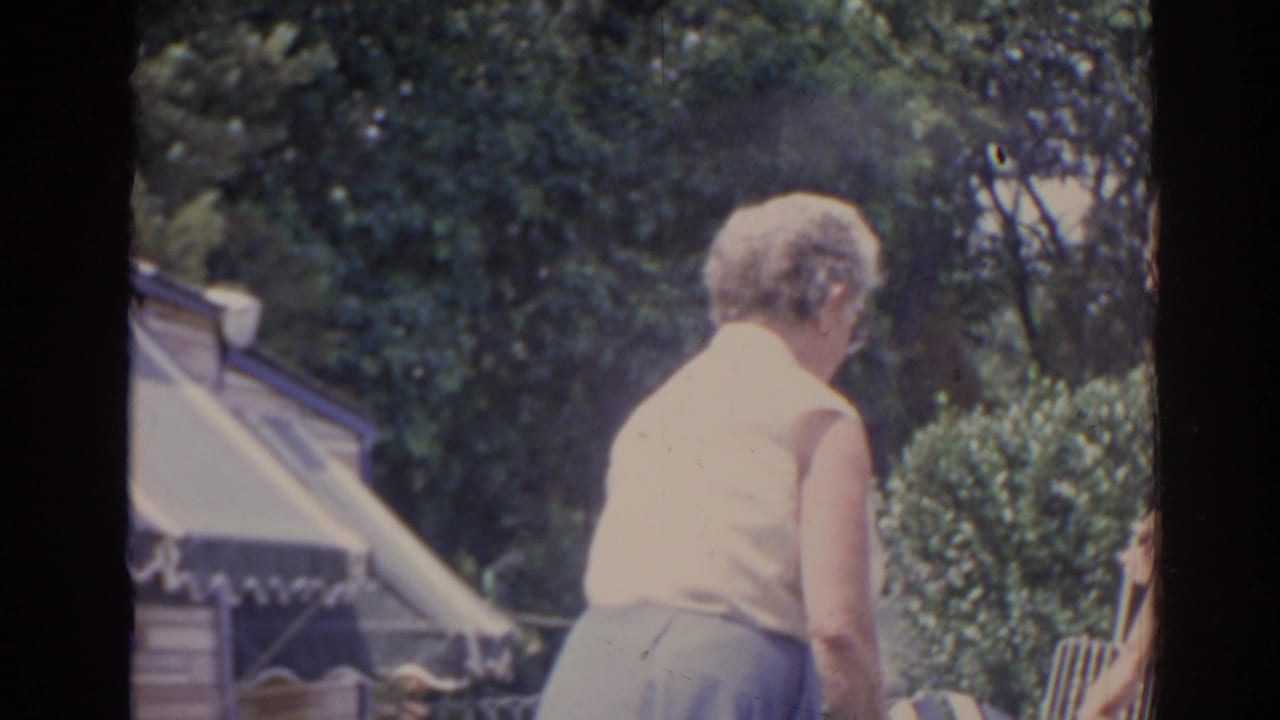Older woman in a sleeveless top stands near a backyard grill with an awning and trees behind her.
