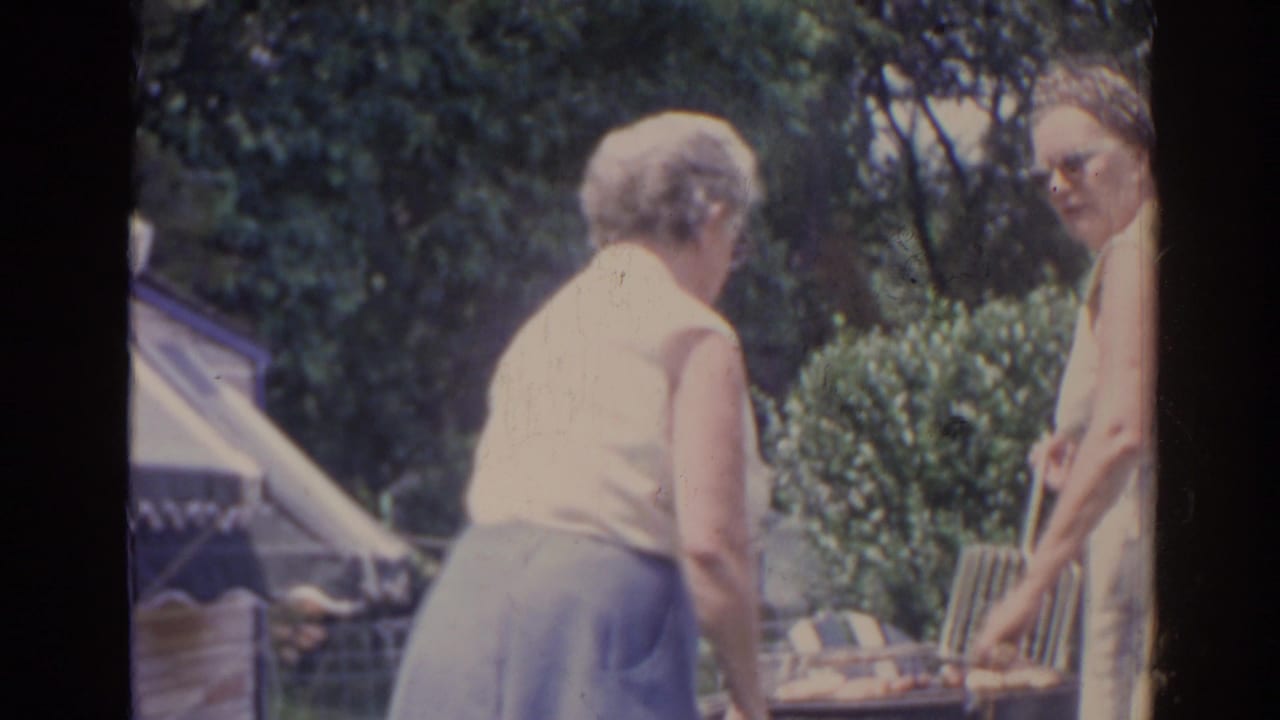 Two older women stand by a backyard grill, with shrubs and a striped awning in the background.