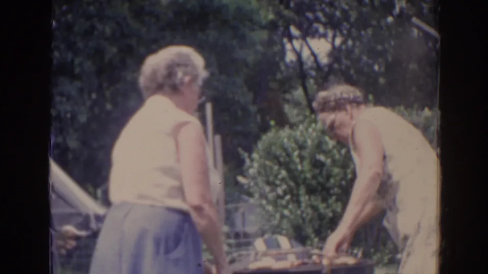 1962 Florida 8mm frame showing one older woman leaning over a backyard grill while another stands near an awning and garden shrubs.