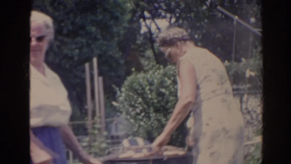 1962 Florida home-movie frame with a woman reaching toward the barbecue grill and another older woman blurred in the foreground.