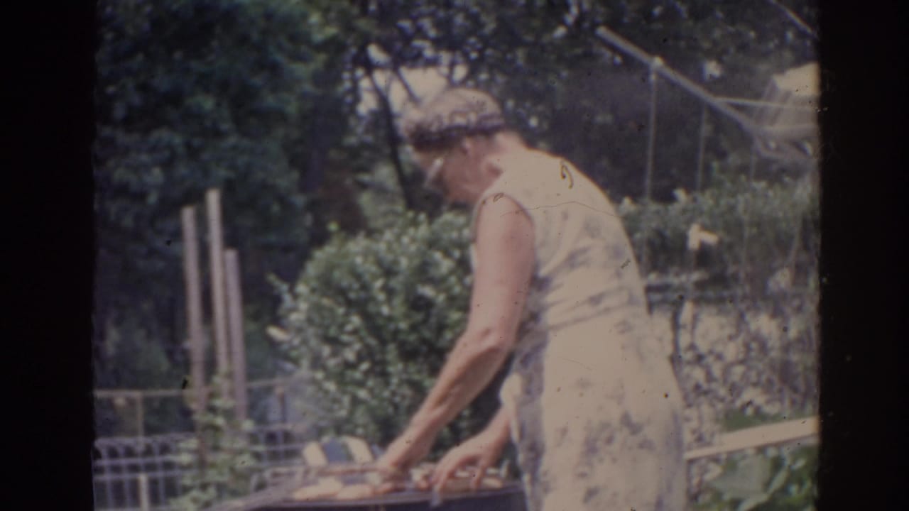 In supported 1962 Florida home-movie footage, an older woman in a light sleeveless dress bends over an outdoor grill in a leafy yard.