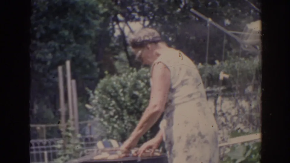 In supported 1962 Florida home-movie footage, an older woman in a light sleeveless dress bends over an outdoor grill in a leafy yard.