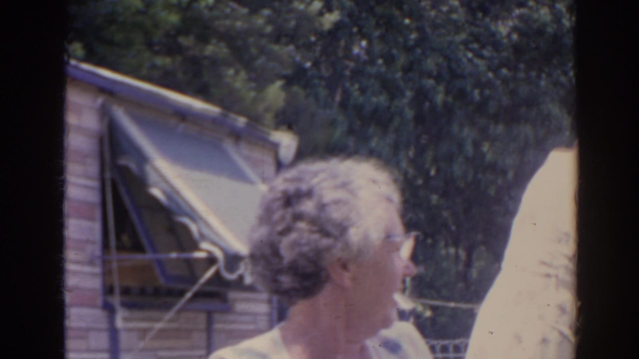 An older woman wearing sunglasses is seen in profile near the striped awning, with trees and the yard behind her.