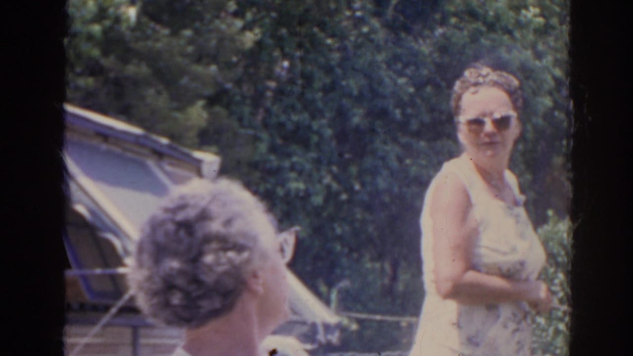Two similarly dressed older women appear in the yard near the striped awning, one turned away and one facing the camera.