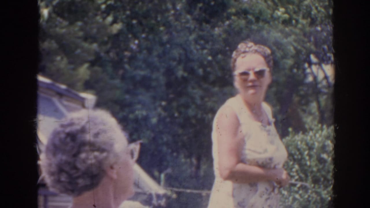 1962 Florida home-movie frame showing adults gathered outdoors, with a woman in sunglasses facing the camera near trees.