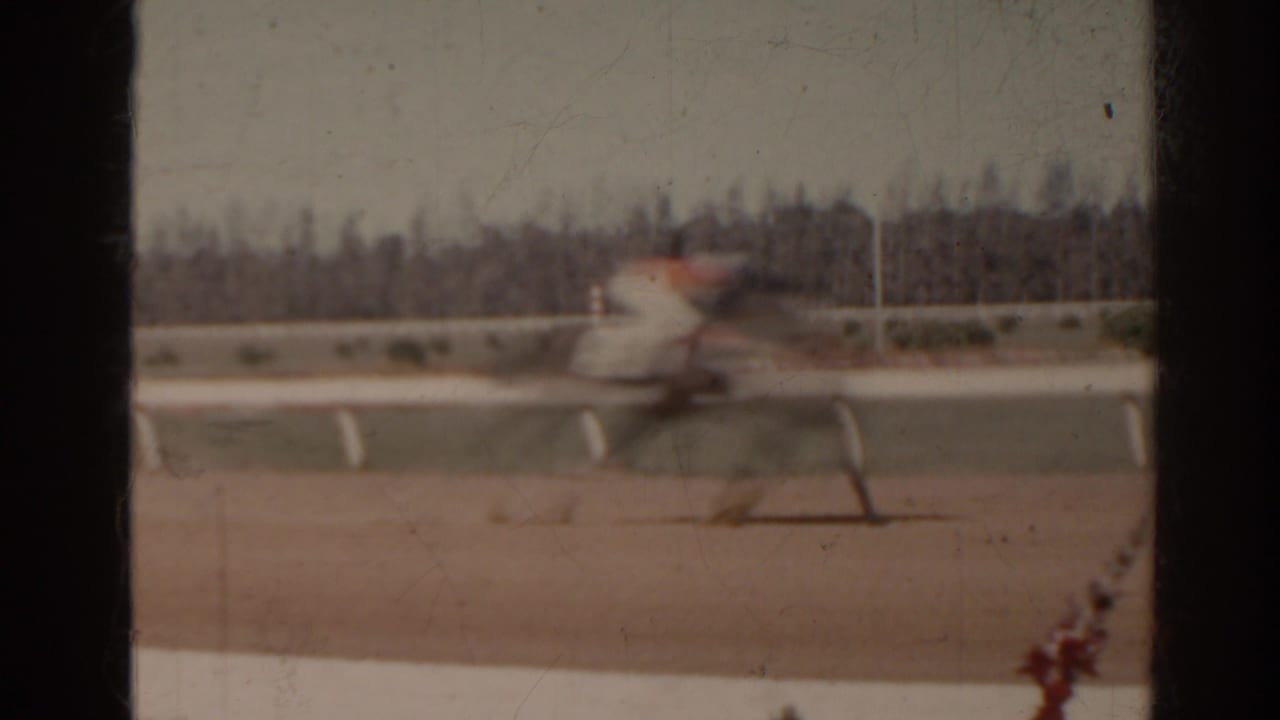 1962 Florida home-movie frame showing a blurred horse and rider moving along a racetrack with white rails.