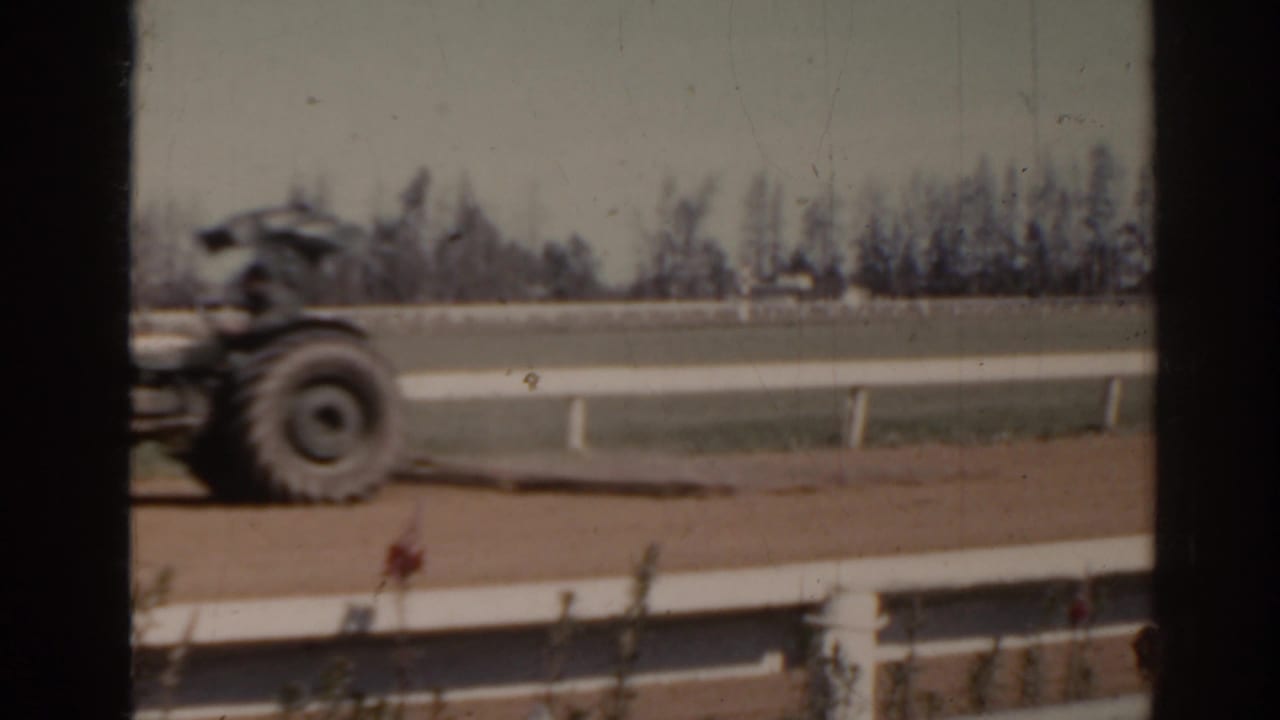 1962 Florida home-movie frame showing a tractor traveling beside a racetrack area with railings and trees beyond.
