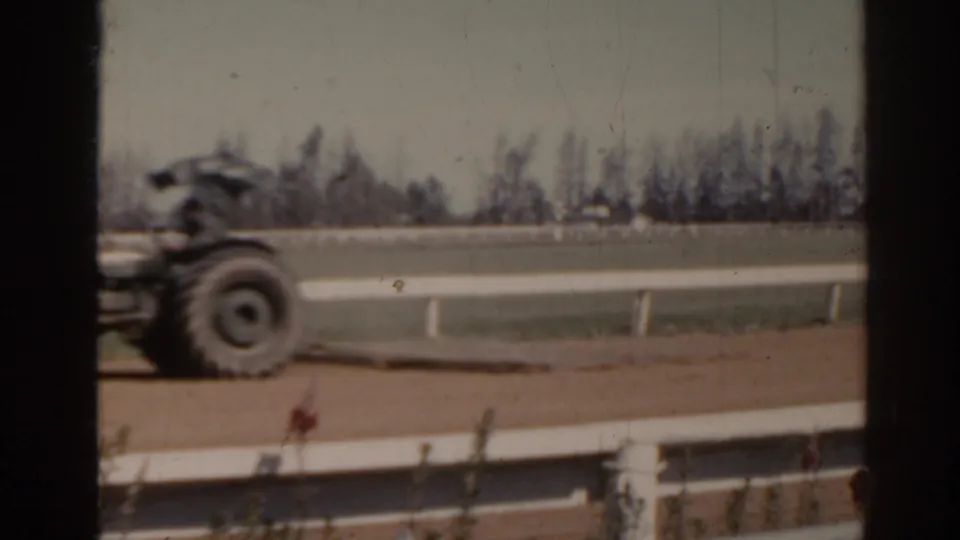 1962 Florida home-movie frame showing a tractor traveling beside a racetrack area with railings and trees beyond.