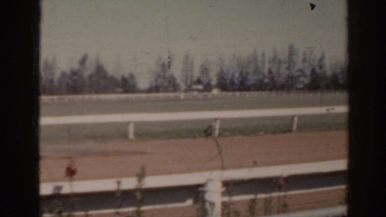 1962 Florida 8mm frame showing an empty horse track with white rails, chain-link fence, distant trees, and pale sky.