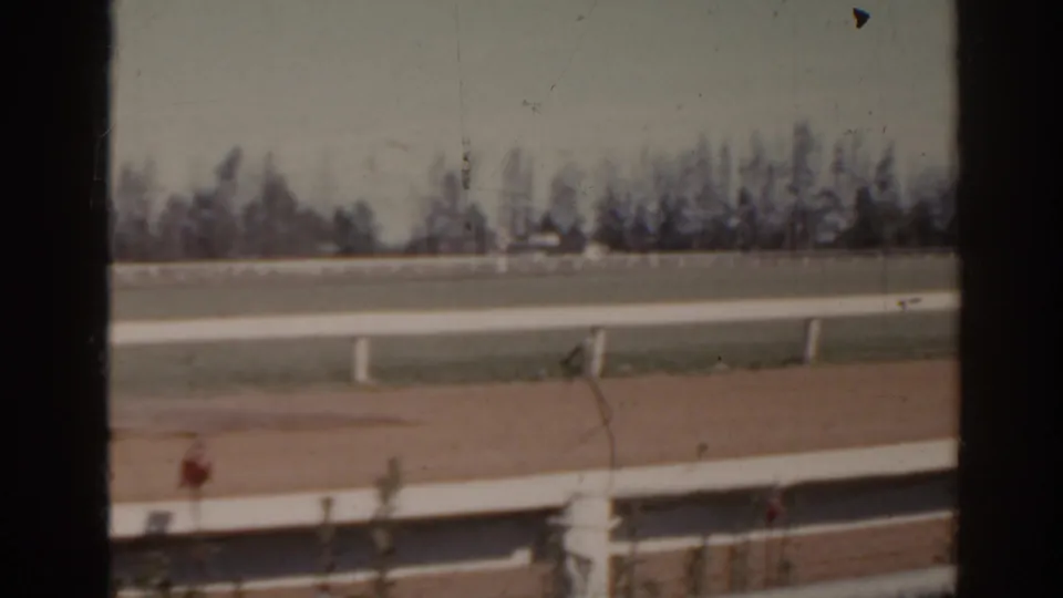 1962 Florida 8mm frame showing an empty horse track with white rails, chain-link fence, distant trees, and pale sky.