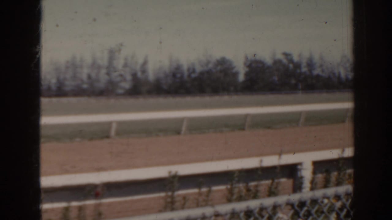 1962 Florida 8mm frame showing the track surface, white railings, chain-link fence foreground, grass, and a soft treeline.