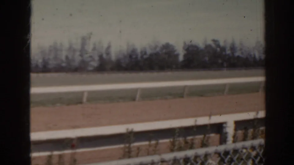 1962 Florida 8mm frame showing the track surface, white railings, chain-link fence foreground, grass, and a soft treeline.