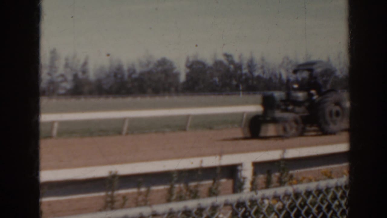 1962 Florida 8mm frame showing a small green tractor moving along the empty dirt horse track near white railings.