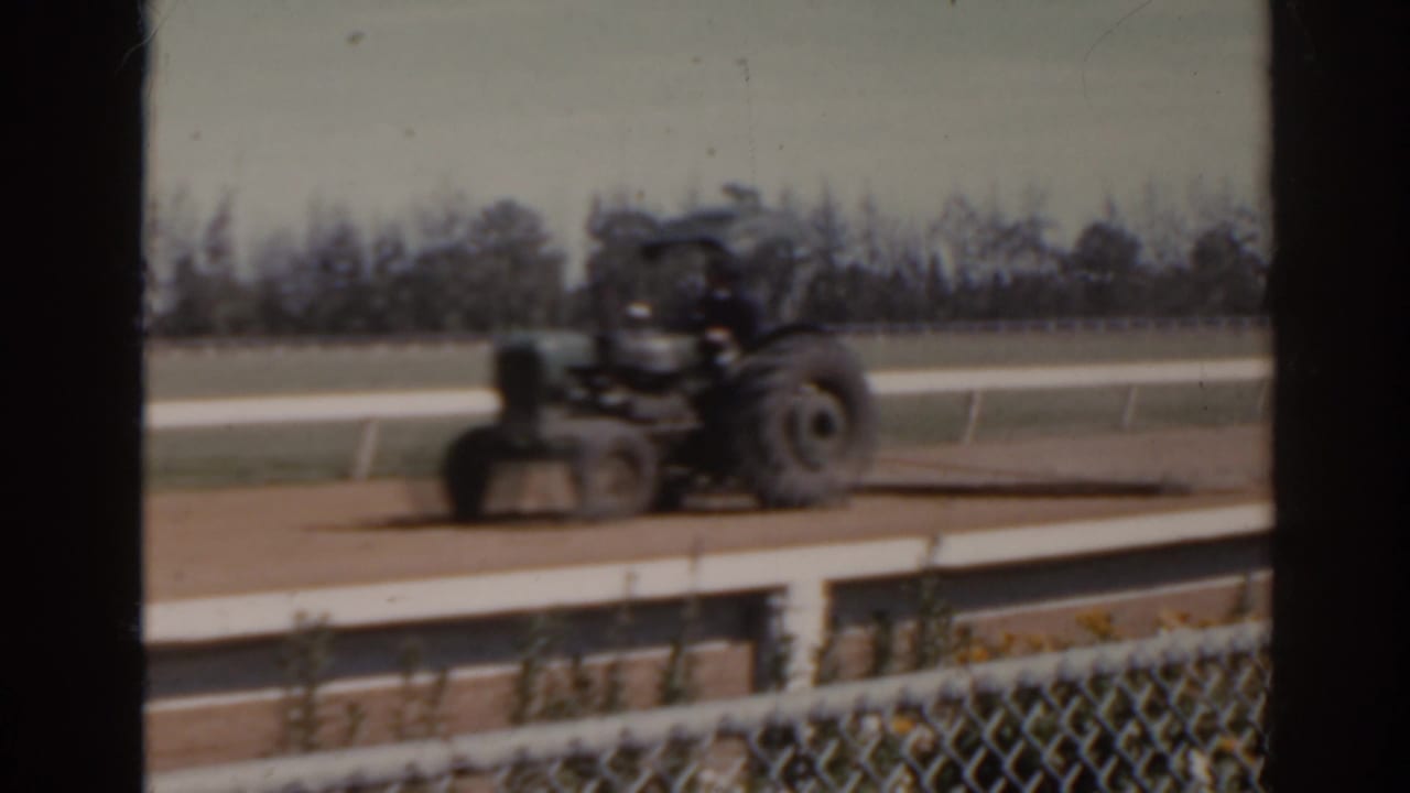 1962 Florida home-movie frame showing a tractor on a dirt racetrack, partly obscured by dark window framing and foreground fencing.