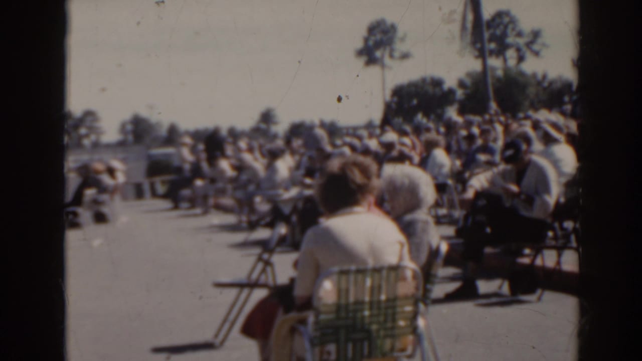 1962 Florida home-movie frame showing spectators in folding chairs near a track, with parked cars and trees in the background.