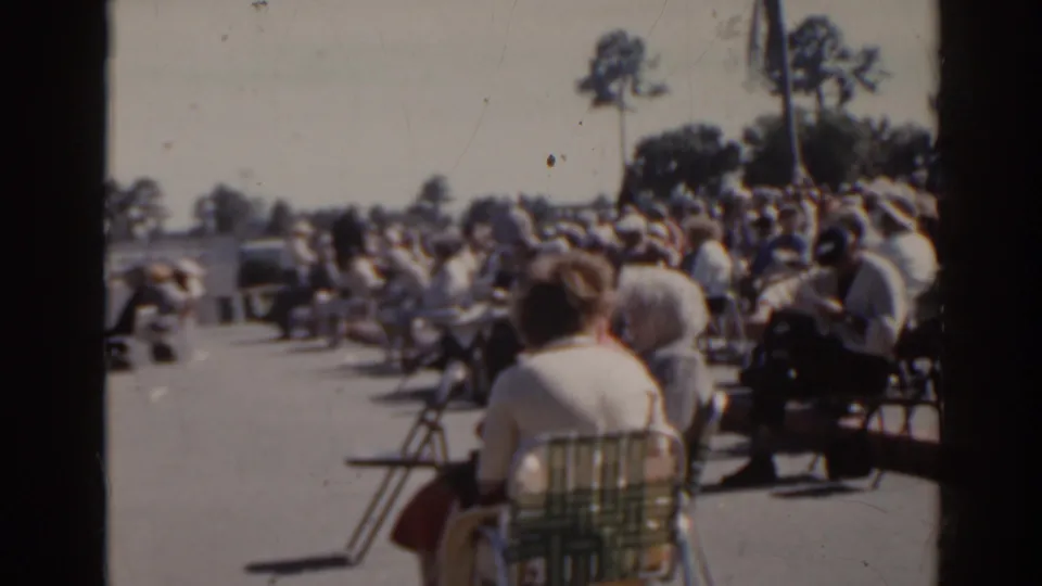1962 Florida home-movie frame showing spectators in folding chairs near a track, with parked cars and trees in the background.