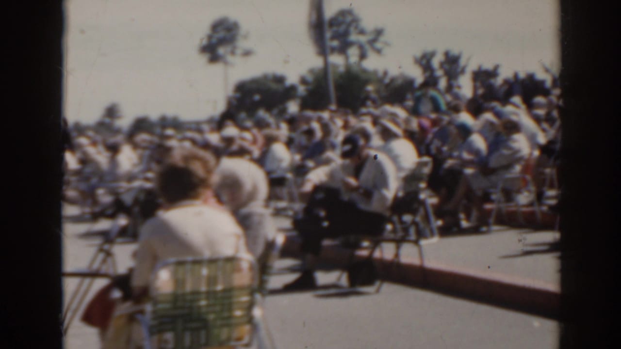 Seated outdoor spectators in folding chairs under bright sun.