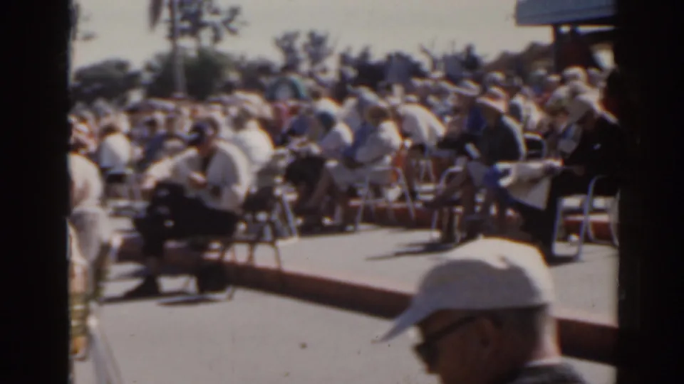 Handheld 8mm view of a 1962 Florida crowd seated in folding chairs, with a raised sign or structure partly visible.