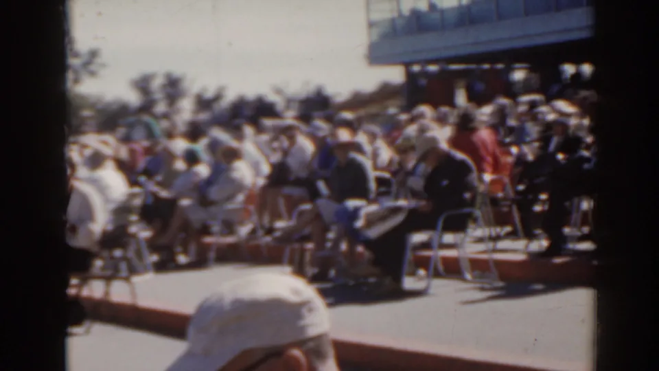 1962 Florida home-movie frame of seated spectators waiting outdoors, with a raised platform or building edge behind them.