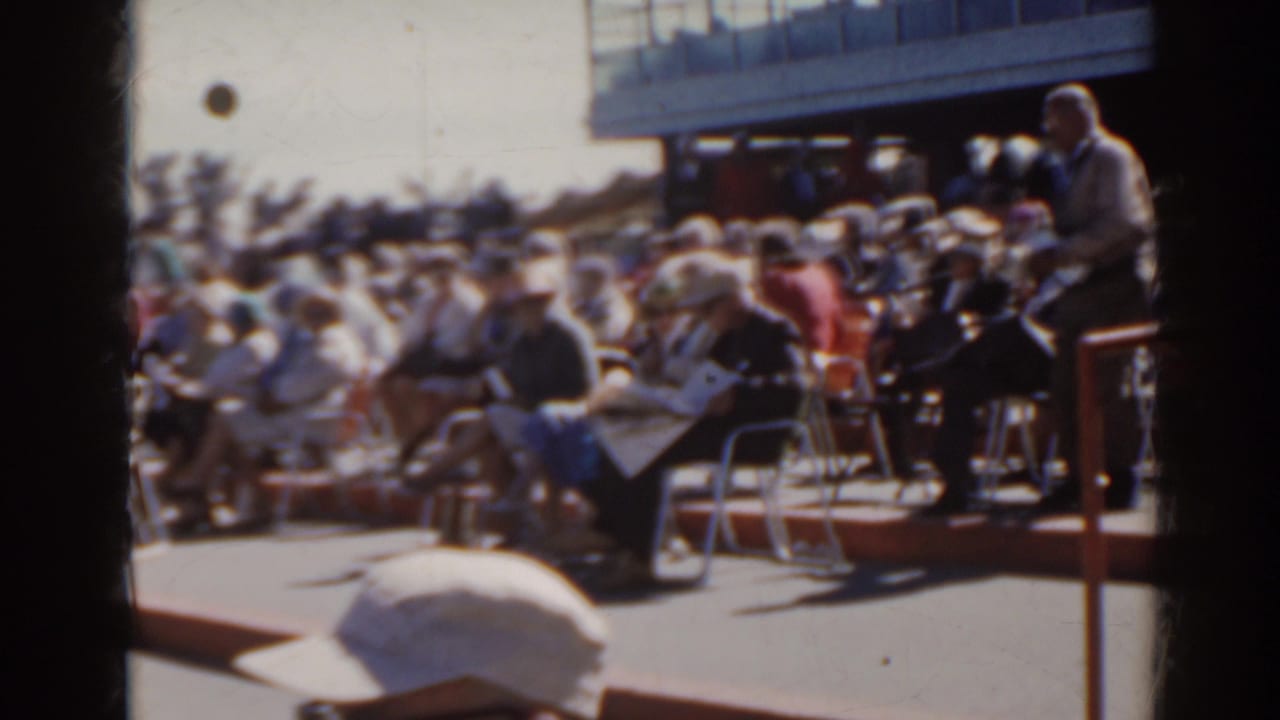 Crowd seated in folding chairs beside a racetrack rail on a bright day.