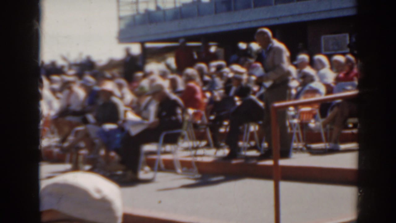 1962 Florida racetrack spectators in outdoor seating, with a suited man standing near the rail.