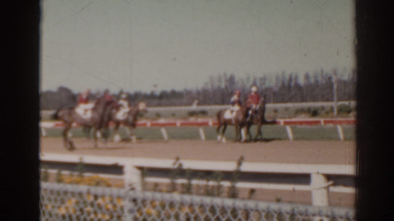 1962 Florida home-movie frame showing horses and riders on a dirt racetrack with spectators out of frame.