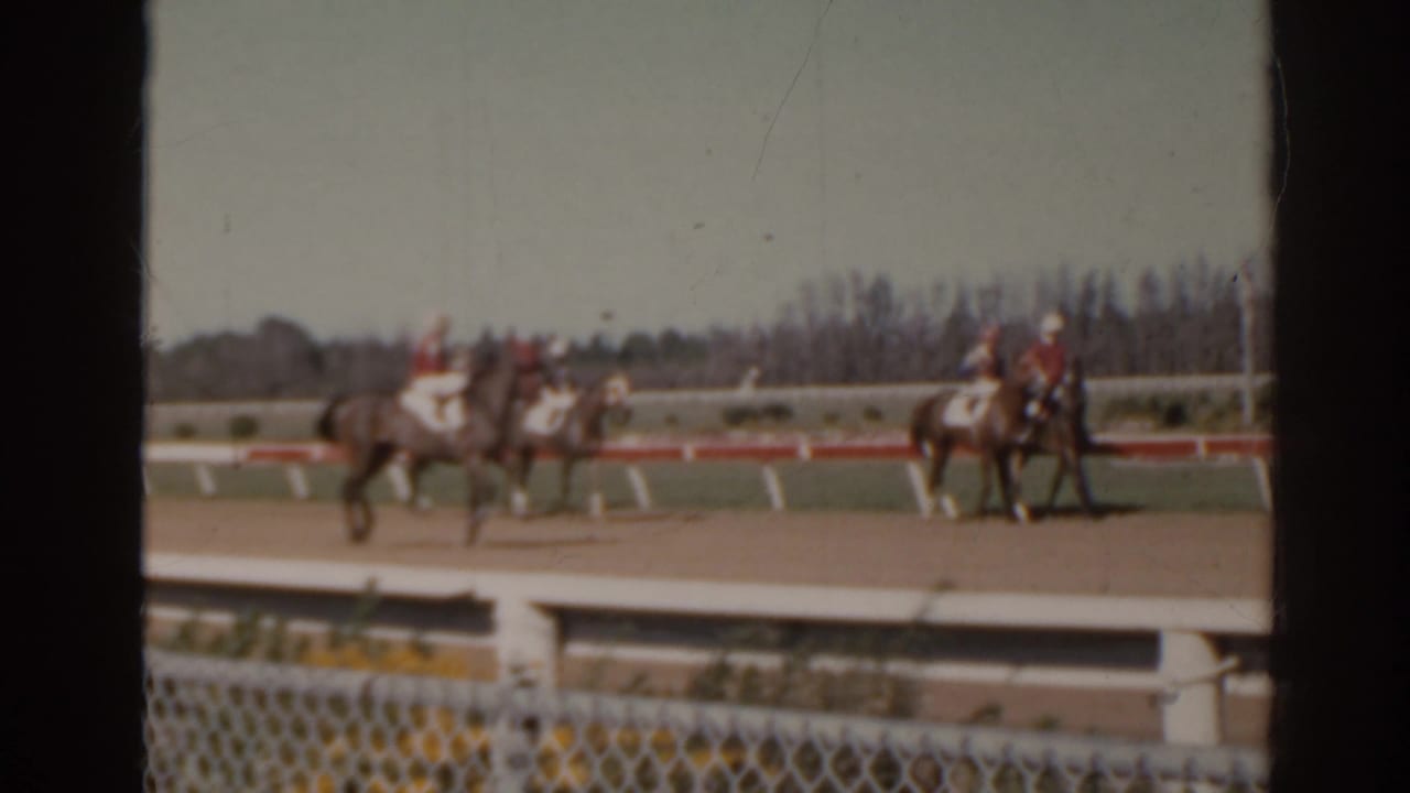 Jockeys on horses move along a dirt racetrack behind white rails and a chain-link fence.