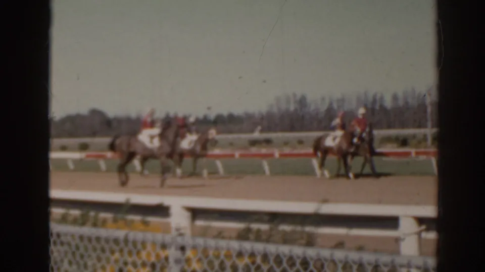 1962 Florida 8mm frame showing jockeys riding horses on a dirt racetrack, seen from behind a chain-link fence.