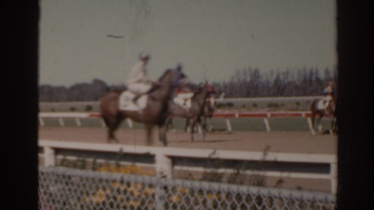 A jockey in light silks rides past on a horse at a Florida dirt track, with more riders and railings in the background.