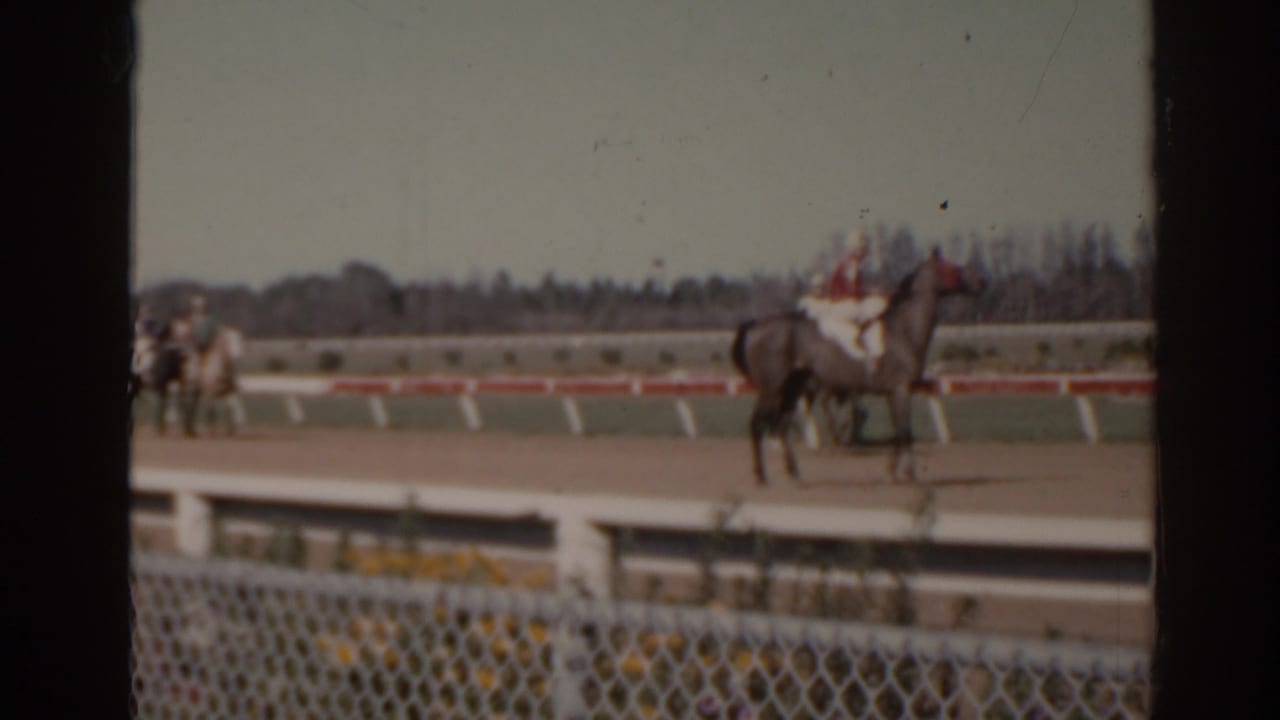 1962 Florida home-movie frame of a jockey in red and white silks on horseback, filmed from behind a foreground fence.