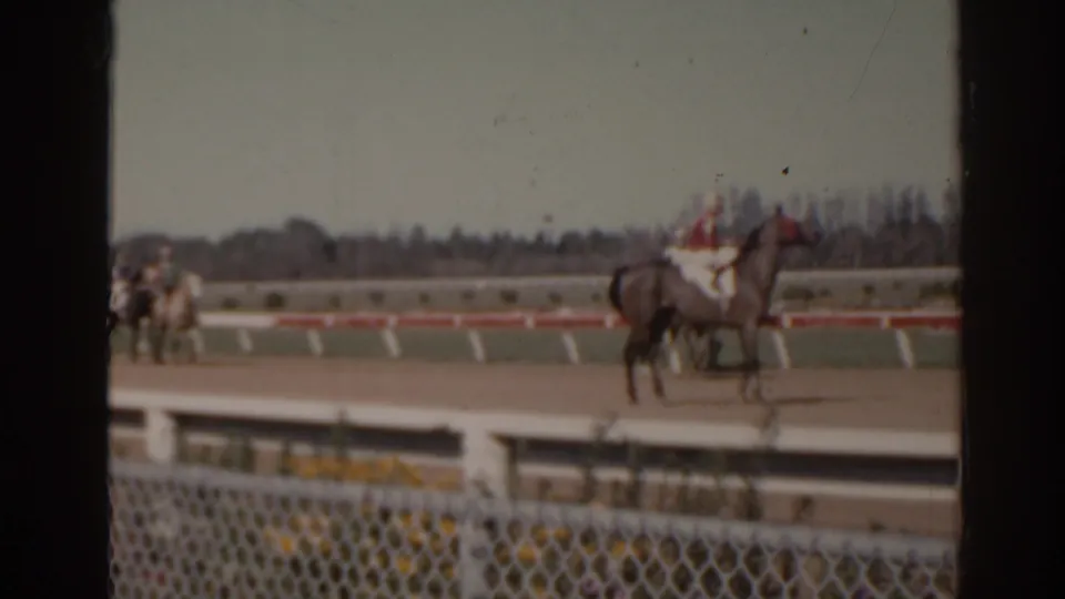 1962 Florida home-movie frame of a jockey in red and white silks on horseback, filmed from behind a foreground fence.